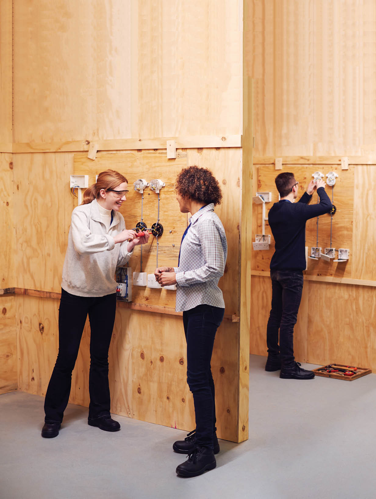 Female Tutor With Trainee Electricians In Workshop Studying For Apprenticeship At College