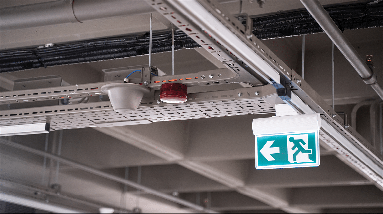 front view closeup of lighted green exit sign hanging on building pipes and pointing the way out of an industrial building