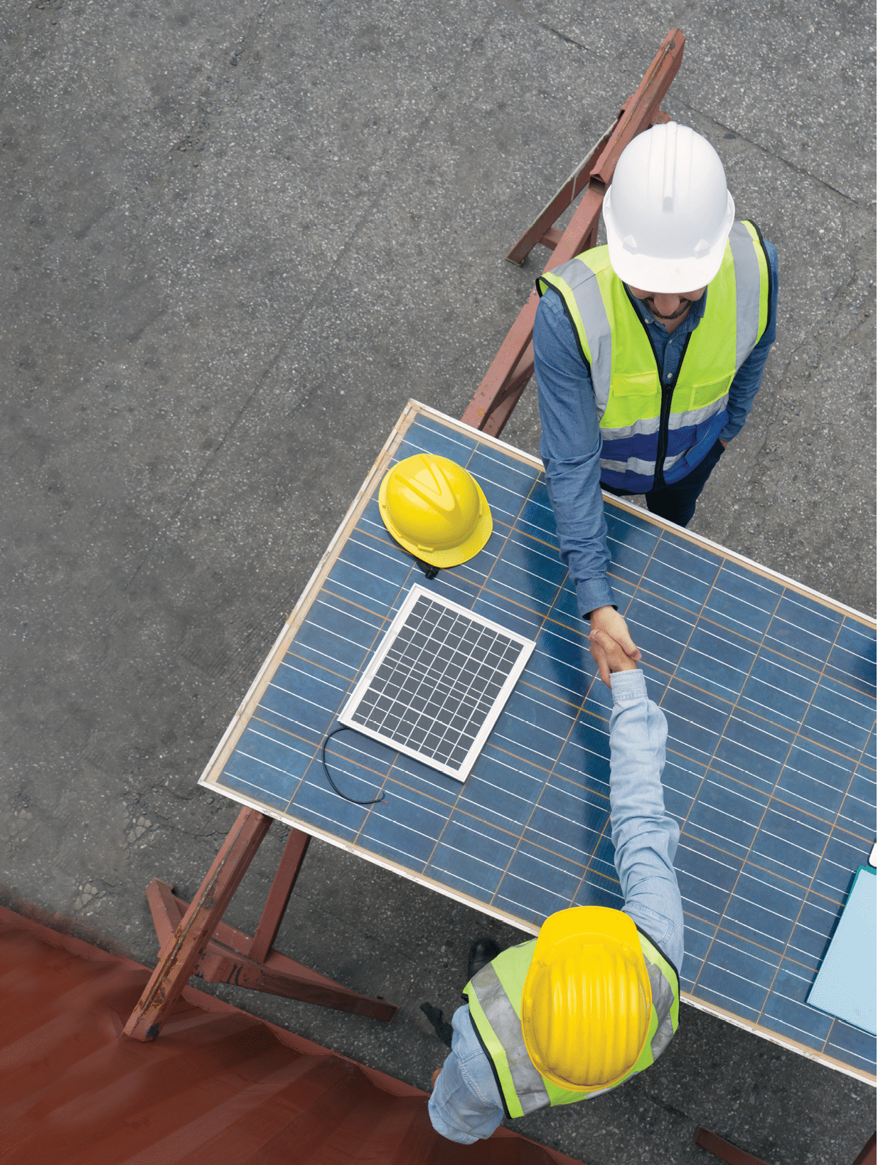 Young engineer in hardhat and safety vest shake hands with project manager after agreeing a business plan  They use solar cell panel as a table  A large container is in the background  Top View
