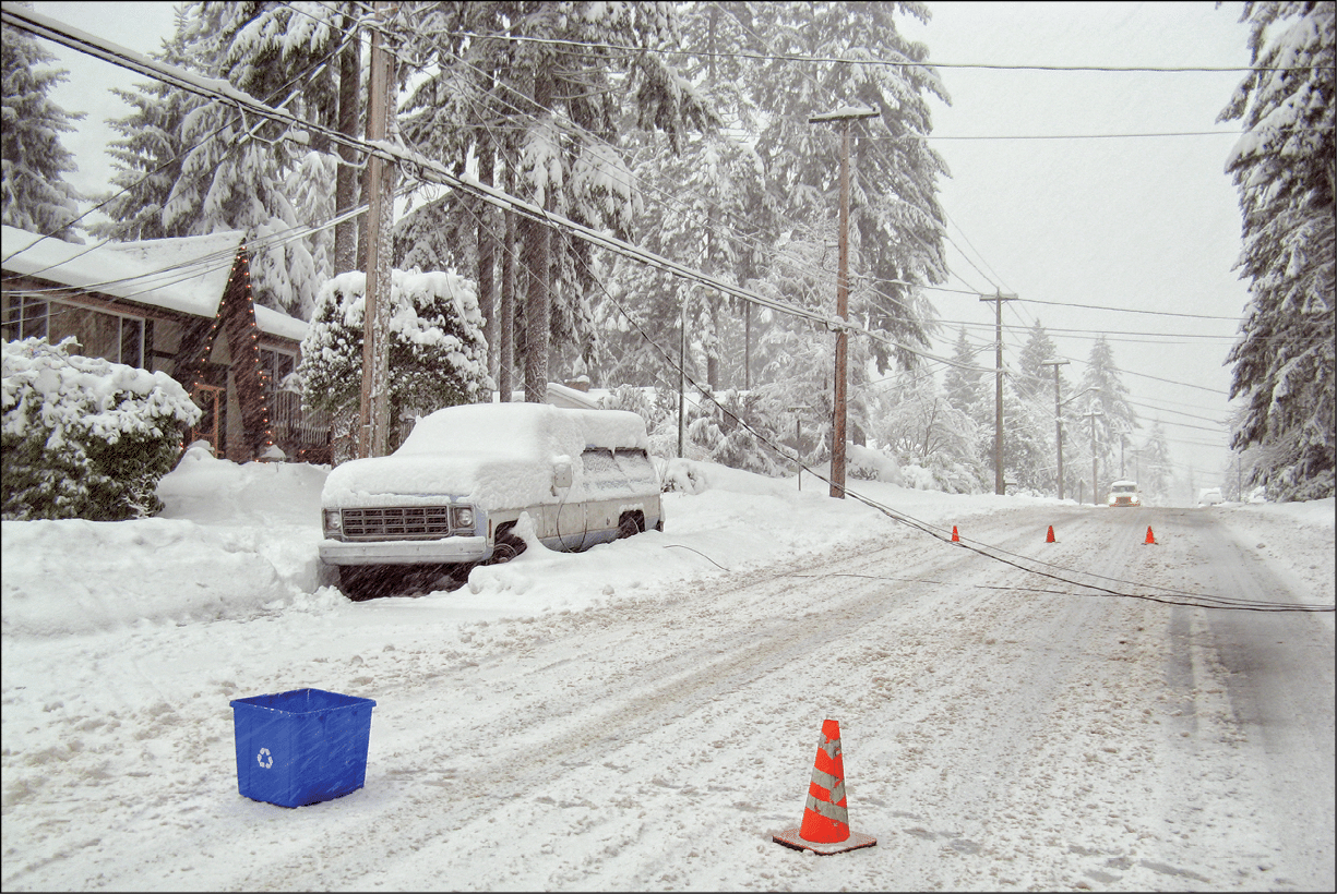 Power lines are on top of a snowed in car and on the snow covered street  A car is approaching  Traffic cones are placed around the area 