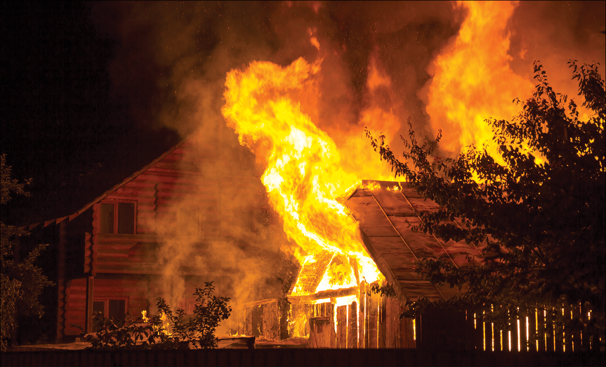 Burning wooden house at night  Bright orange flames and dense smoke from under the tiled roof on dark sky, trees silhouettes and residential neighbor cottage background  Disaster and danger concept 