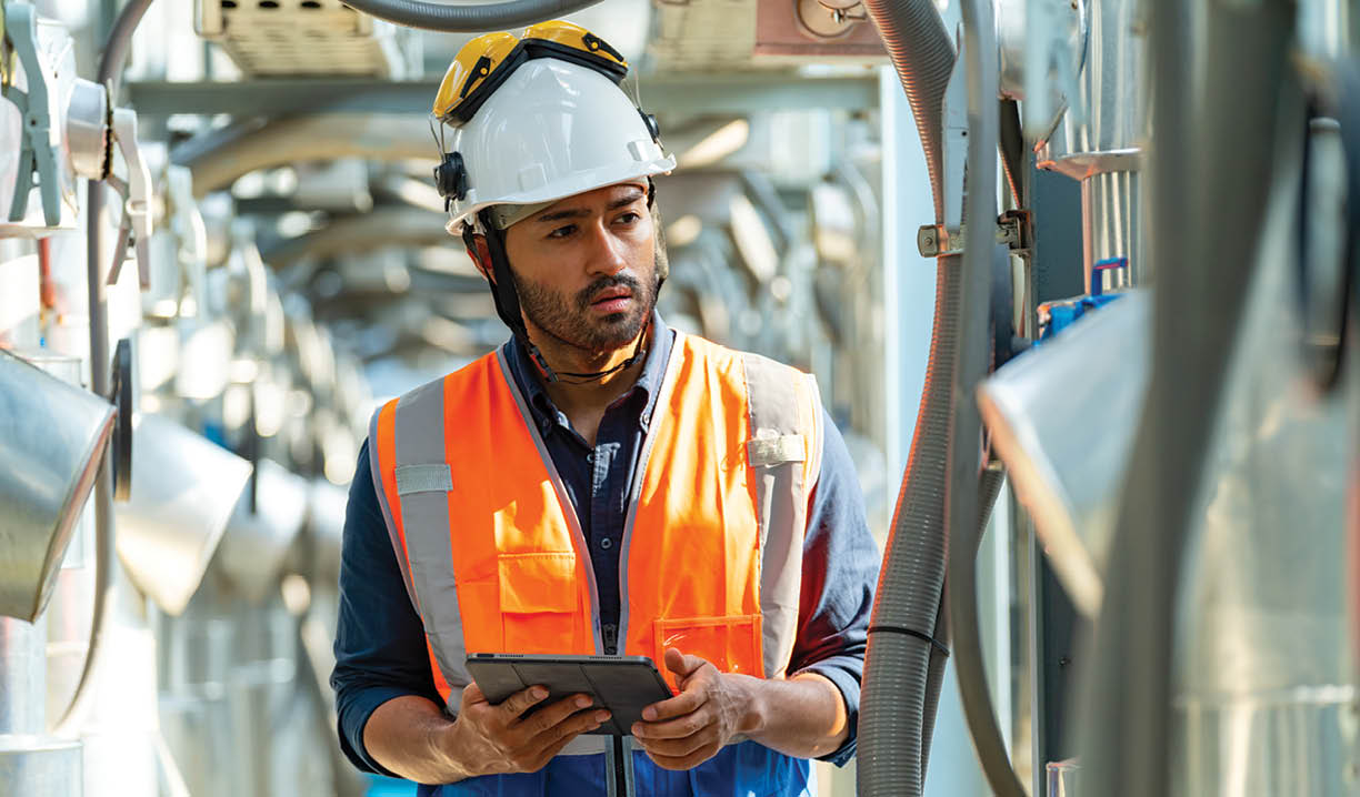 Professional Asian man engineer in safety uniform working on digital tablet at outdoor construction site rooftop. Industrial technician worker maintenance checking building exterior air HVAC systems.