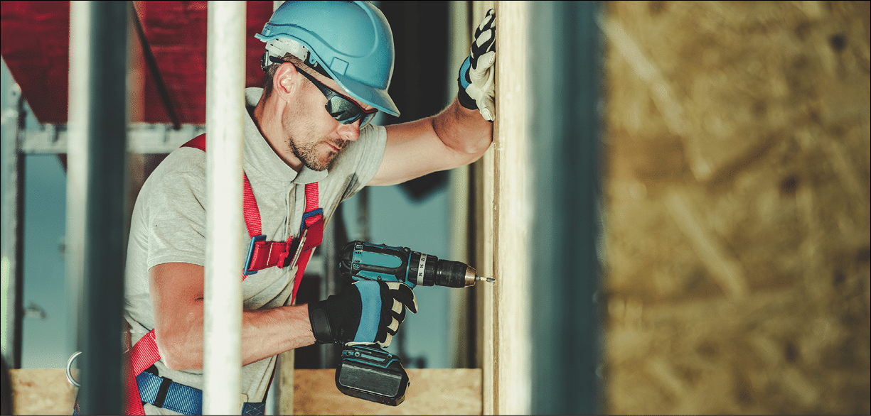 Construction at Height Job. Caucasian Contractor Wearing Hard Hat with Drill Driver Attached by Safety Harness to a Scaffolding.
