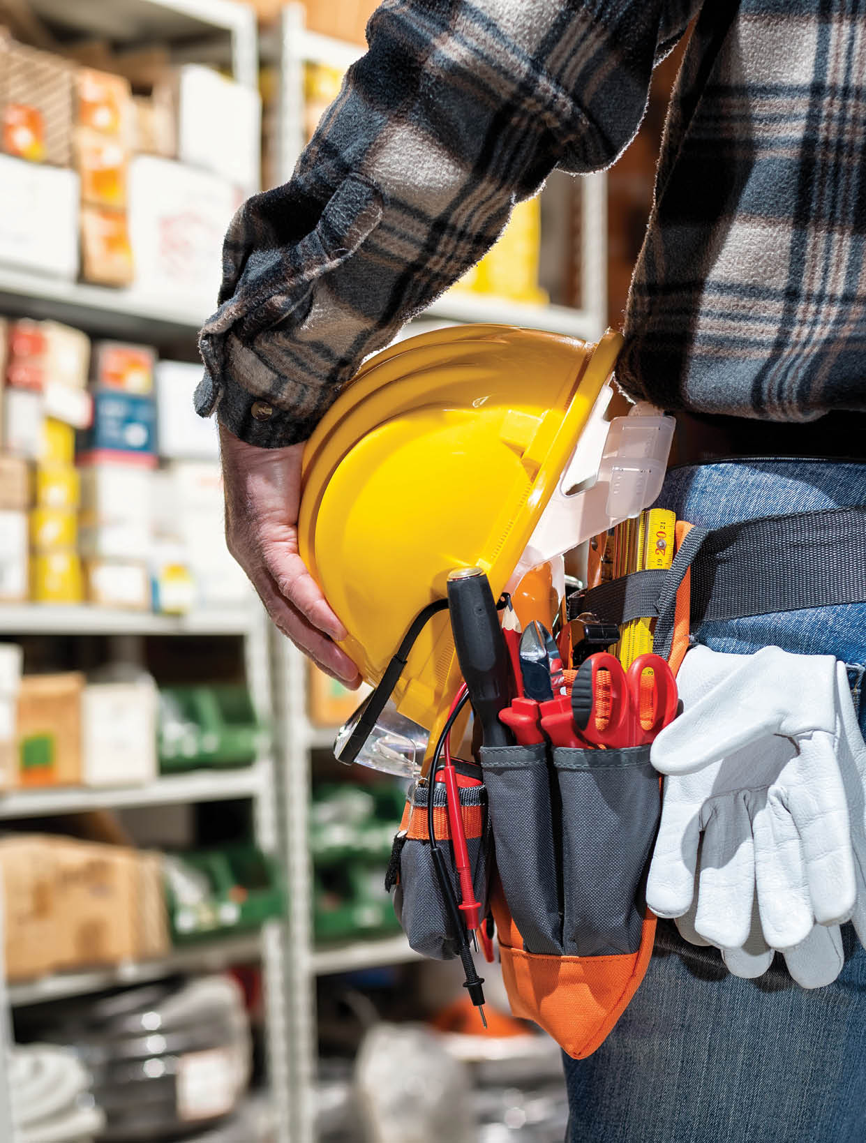 Electrician in the electrical component store holds the helmet and goggles in his hand. Construction industry, electrical system. View from behind.