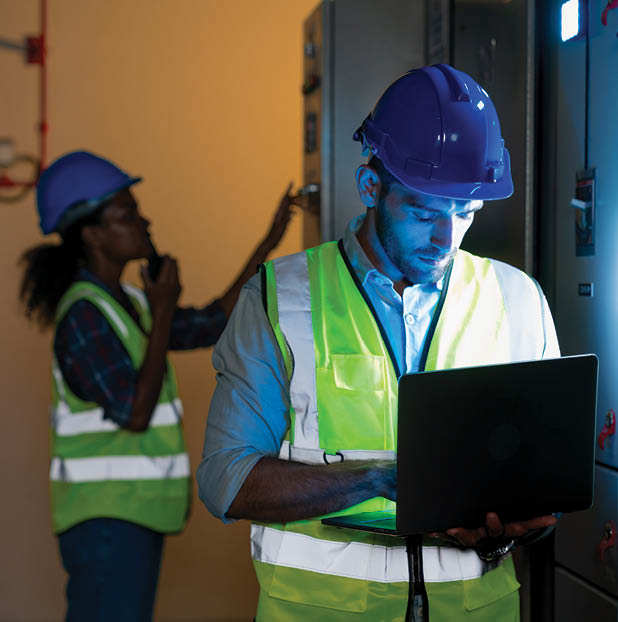 Electrical engineer use laptop to inspection and testing of electrical system in control room. Electrician and technician working night shifts at distribution board. Multi-Ethnic Blue-collar worker.