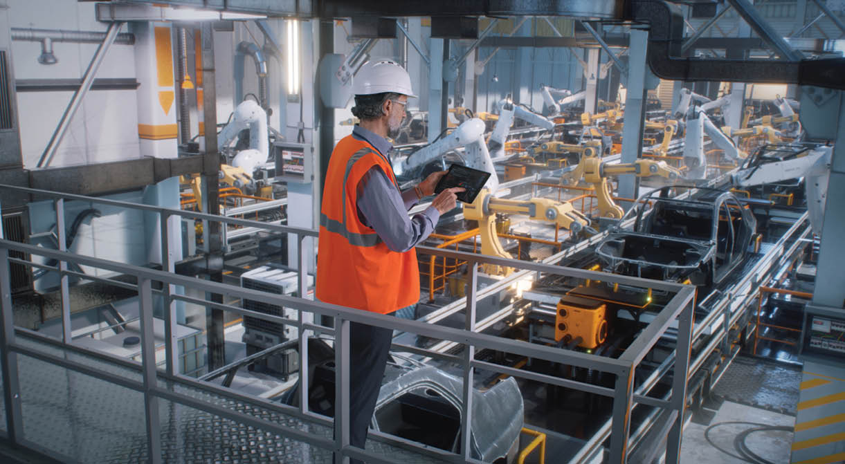 High angle of mature male engineer in uniform and hardhat using tablet to control robotic arms during work on contemporary car factory