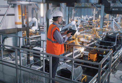 High angle of mature male engineer in uniform and hardhat using tablet to control robotic arms during work on contemporary car factory