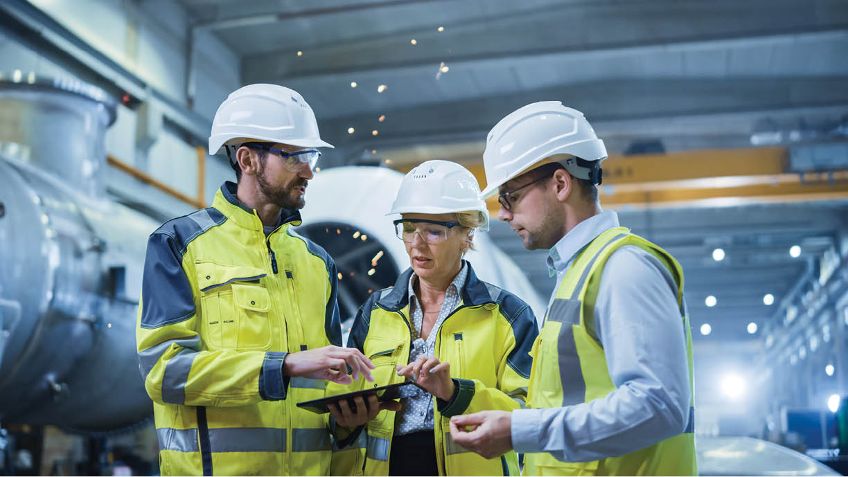 Three Heavy Industry Engineers Stand in Pipe Manufacturing Factory, Use Digital Tablet Computer, Have Discussion. Large Pipe Assembly. Design and Construction of Oil, Gas and Fuels Transport Pipeline